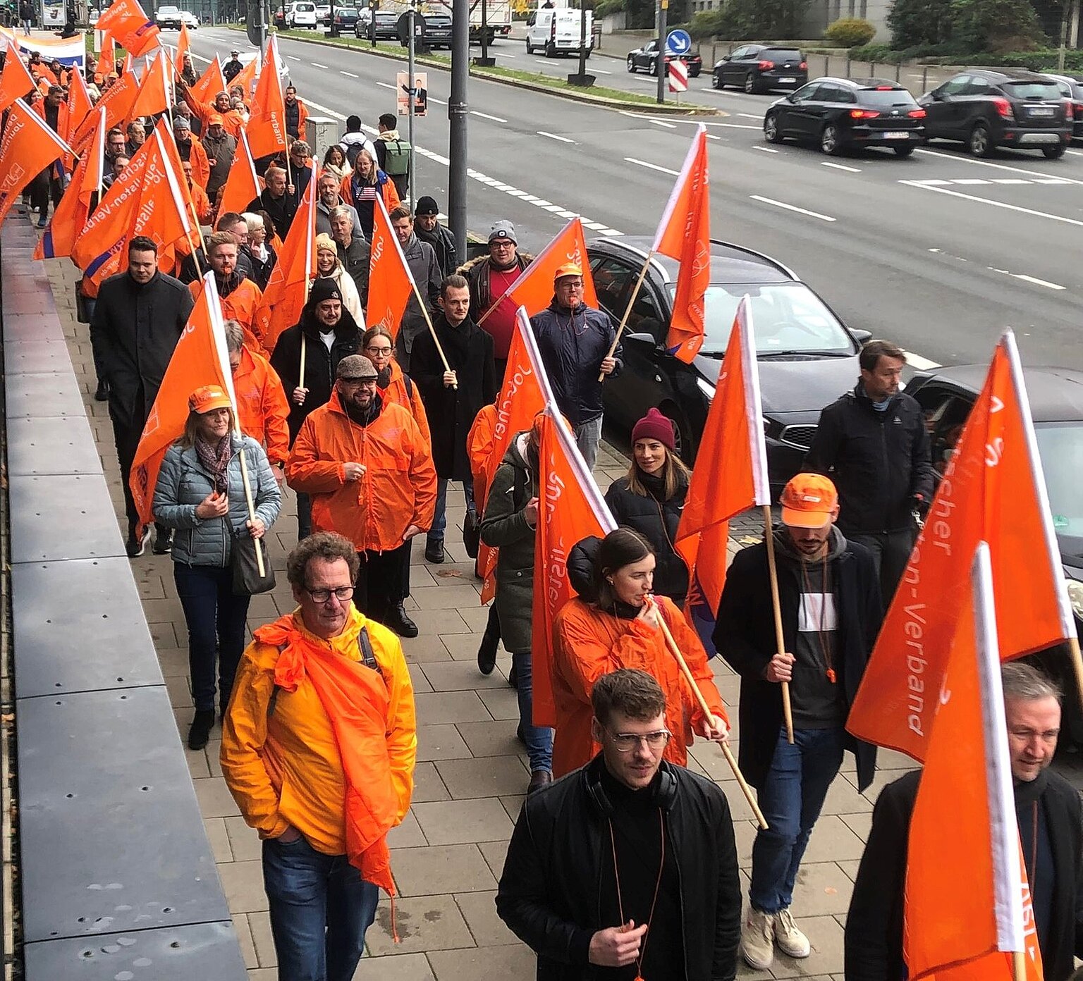 Ein Streikzug aus Fahnenträgern in orangenen Jacken. – Demonstration vor der Funke-Zentrale in Essen. Foto: Michael Kleinrensing