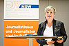 Andrea Hansen steht am Stehtisch und spricht, im Hintergrund ein Banner des DJV-NRW. – 