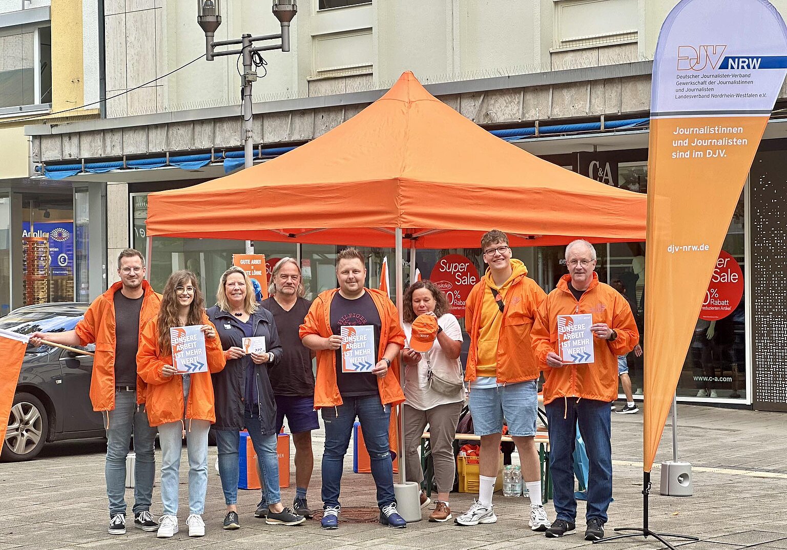 Eine Gruppe von Personen steht vor einen orangenen Pavillion, sie tragen orangene Jacken und halten Streikschilder in die Kamera. – 